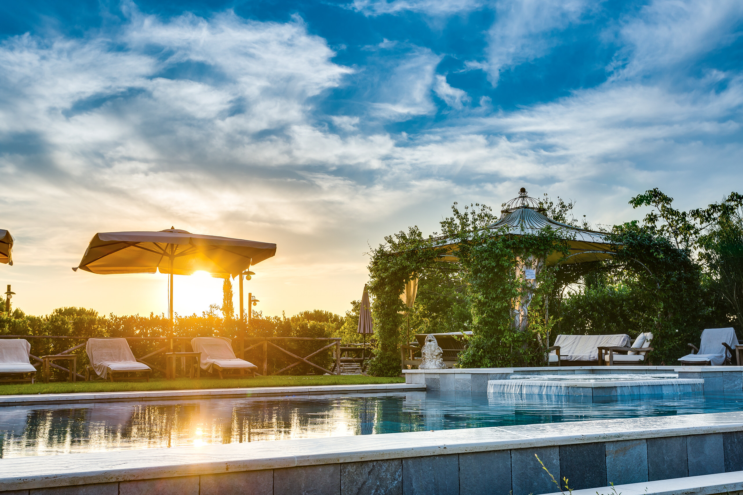 una piscina al tramonto con un gazebo a cupola sullo sfondo, ombrellone e sedute a bordo vasca, sole basso che si riflette sull'acqua.