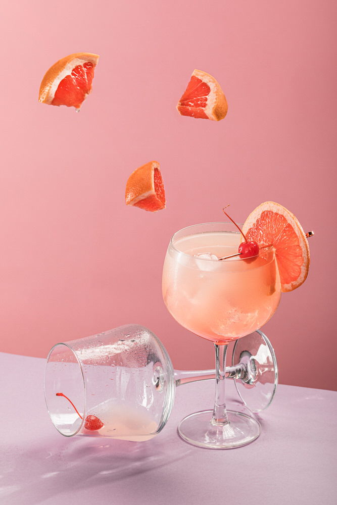 Cocktail rosato in coppa con pompelmo e ciliegia, spicchi sospesi in aria su fondo rosa