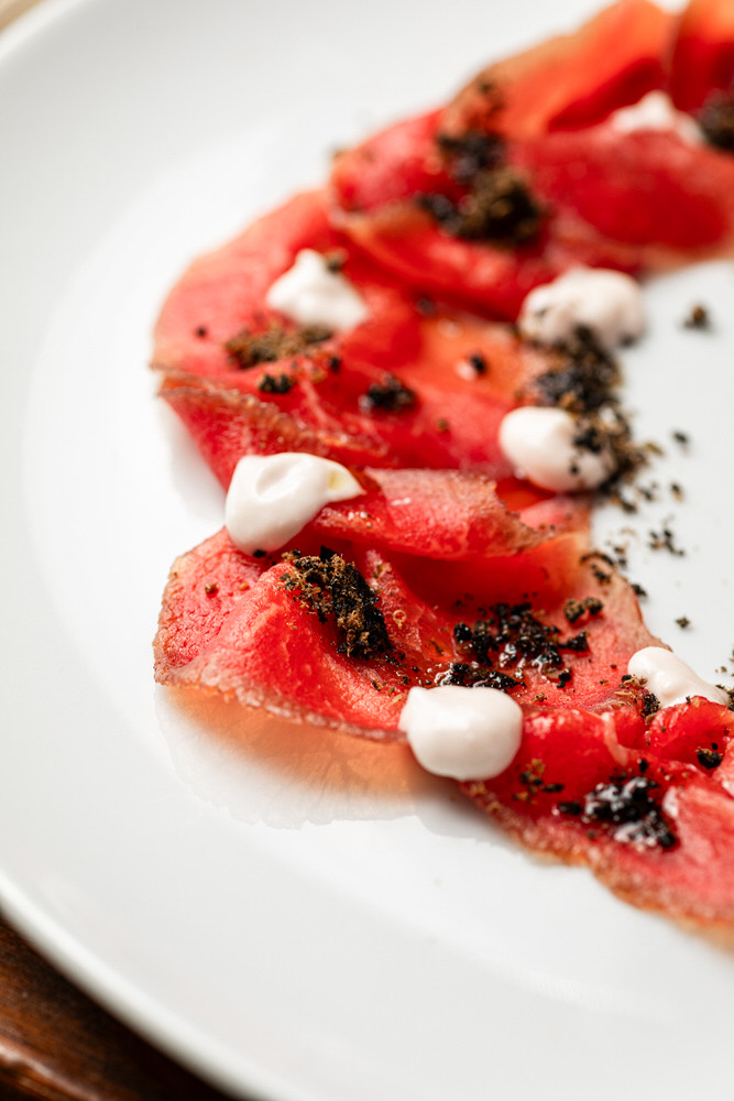 Dettaglio ravvicinato di un carpaccio di carne con crema e crumble scuro su piatto bianco